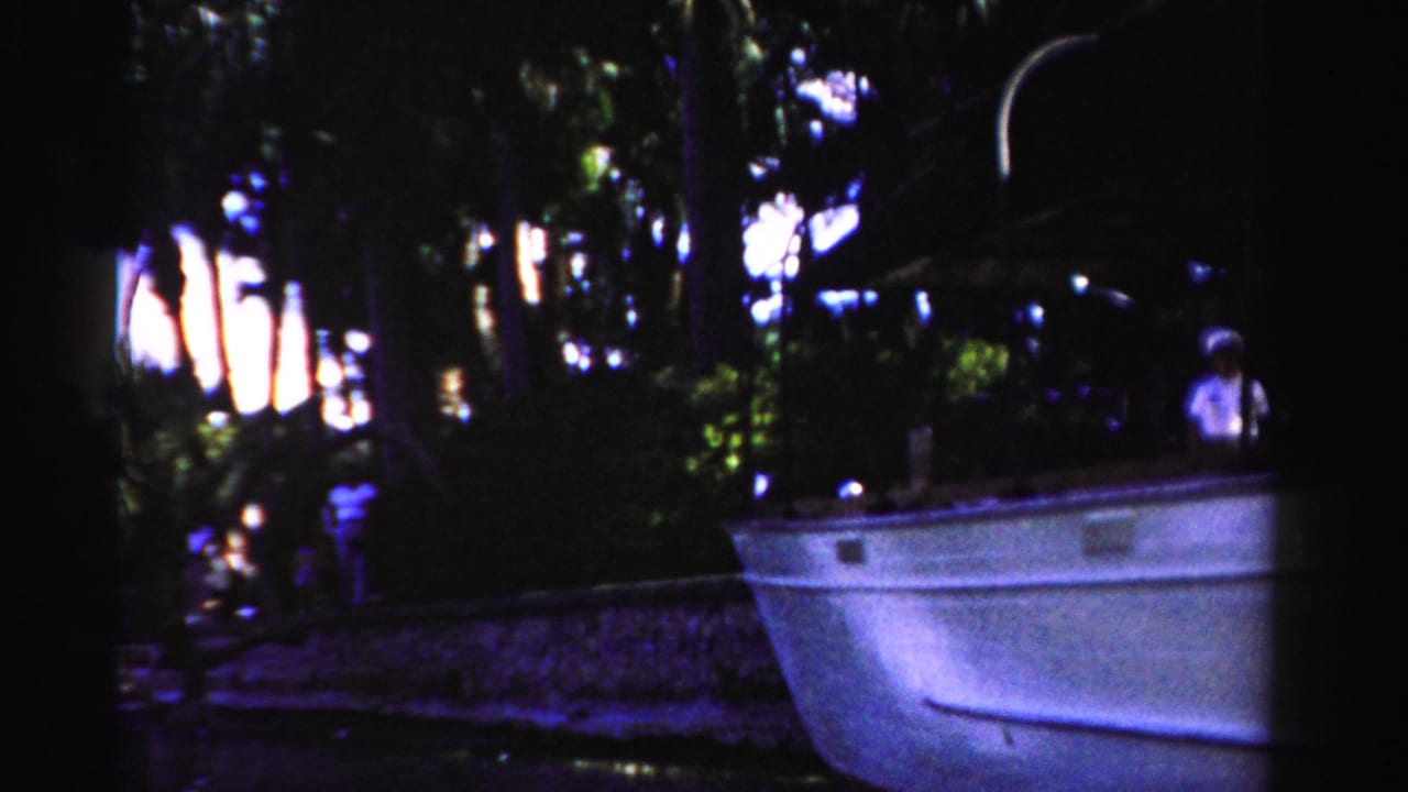 Small boat beside a dark wooded riverbank with a person standing nearby.