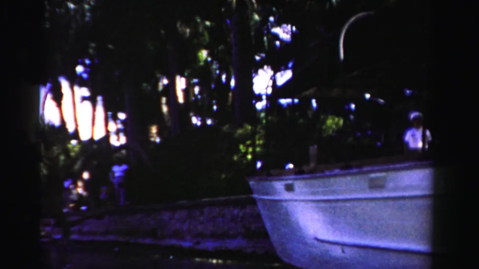 1955 Dade County 8mm frame showing a small boat at a shaded riverside edge with a person in a light shirt near the stern.
