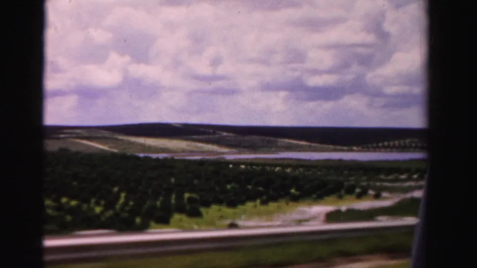 1955 Dade County landscape frame showing cultivated rows, a winding road, low distant horizon, and clouded sky from a moving viewpoint.