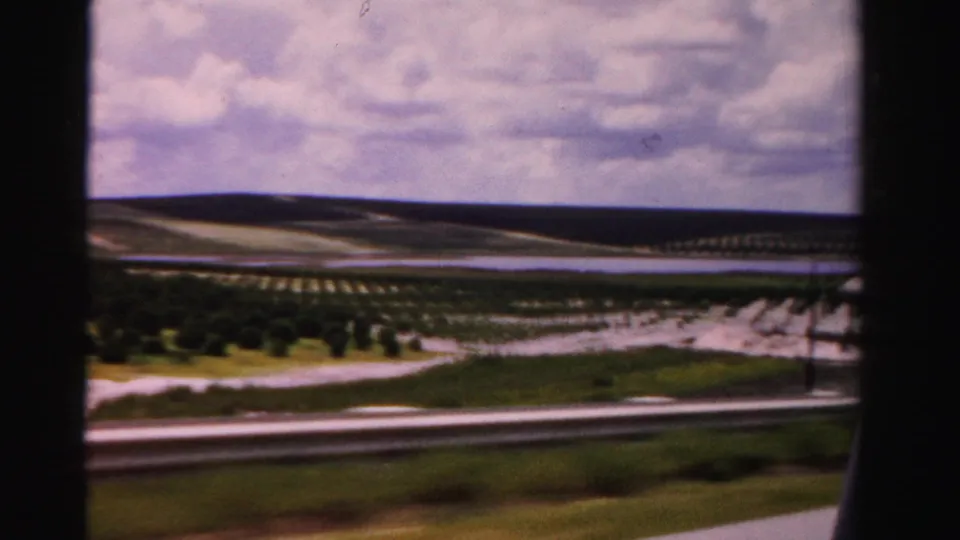 Moving-vehicle view in 1955 Dade County, with a guardrail, planted rows, open fields, and soft 8mm color.