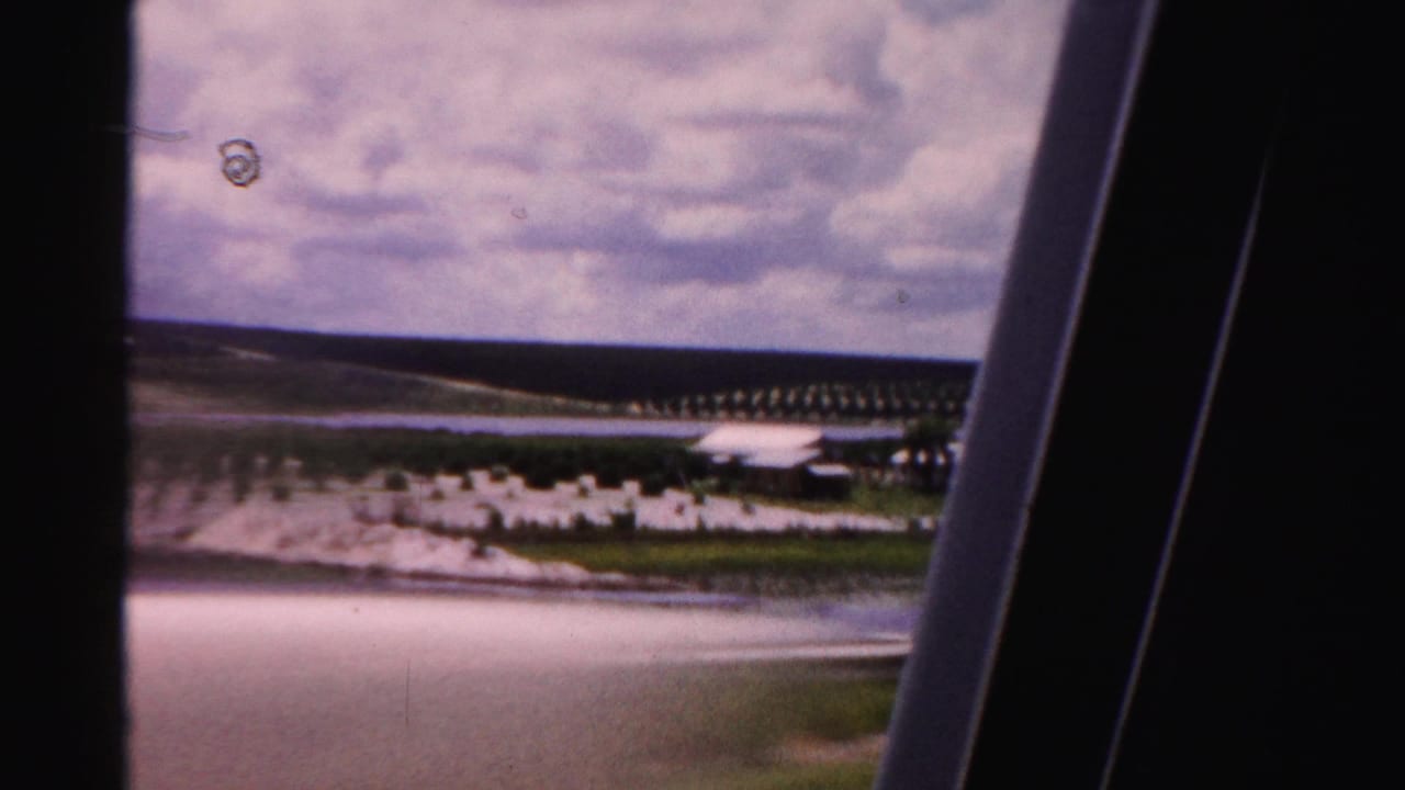 Agricultural rows, water, low white buildings, cloudy sky, and vertical car-window edges.