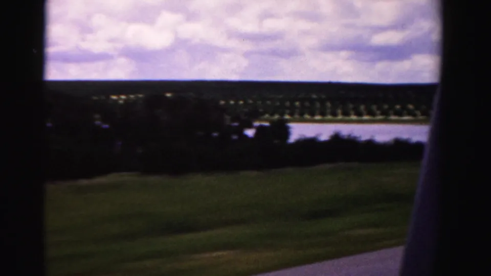 Dade County 8mm travel frame with motion-blurred roadside grass, distant planted rows, and water under a cloudy sky.