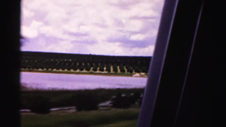 Moving-vehicle 8mm frame showing tree rows along water under a cloudy sky in Dade County, Florida.