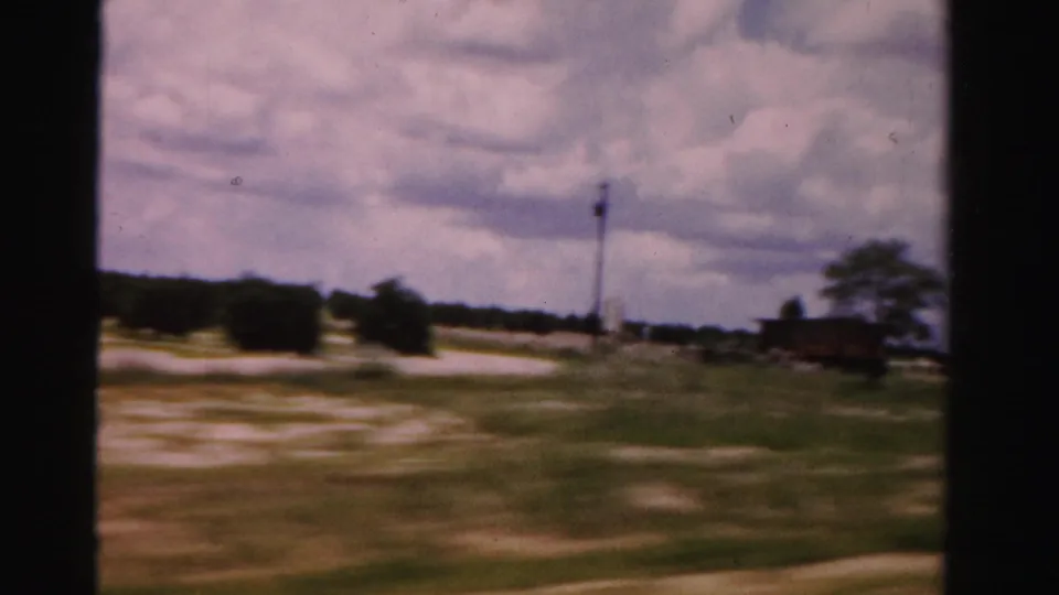 Dade County road footage shows open sandy roadside ground, a utility pole, distant trees, and cloudy sky from a moving car.