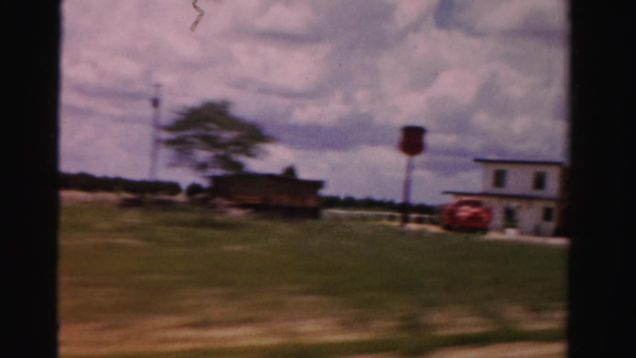 1955 Dade County road-trip frame showing open grass, a low roadside building, a red car, trees, and a distant tower from a moving vehicle.