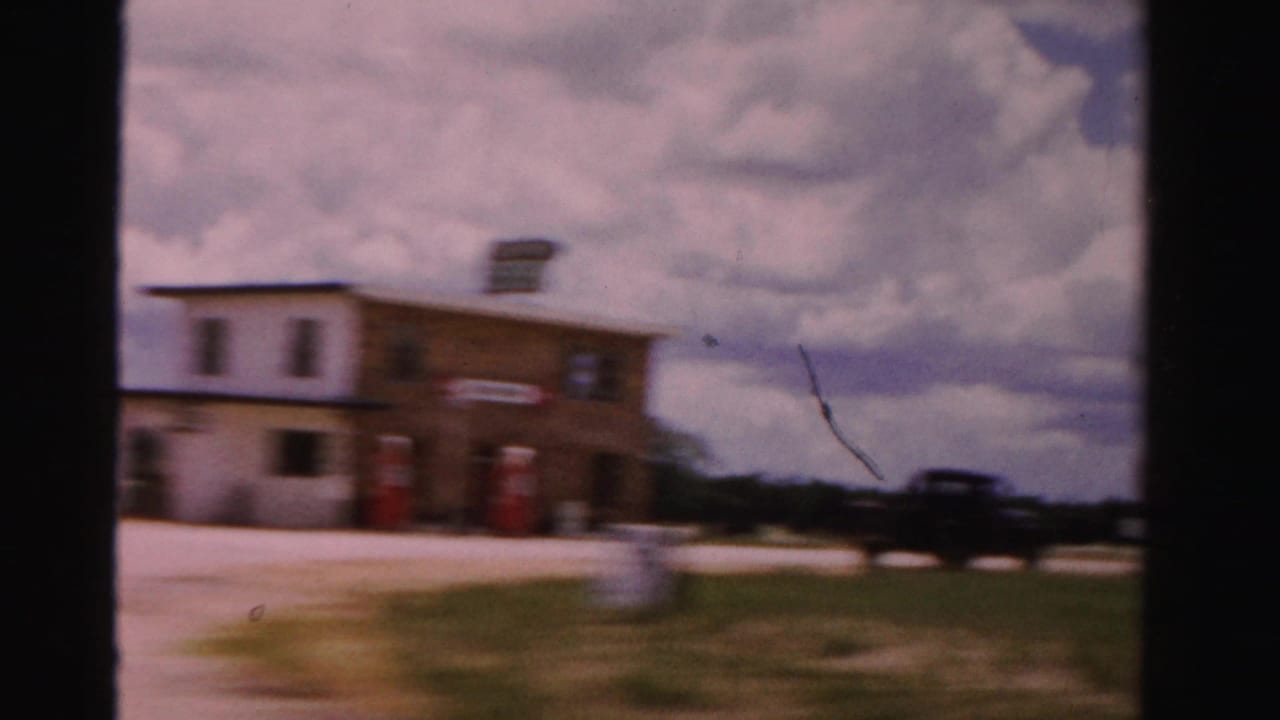 1955 Dade County roadside gas station scene with red pumps, a low building, parked cars, grass, and a cloudy sky, softened by motion blur.