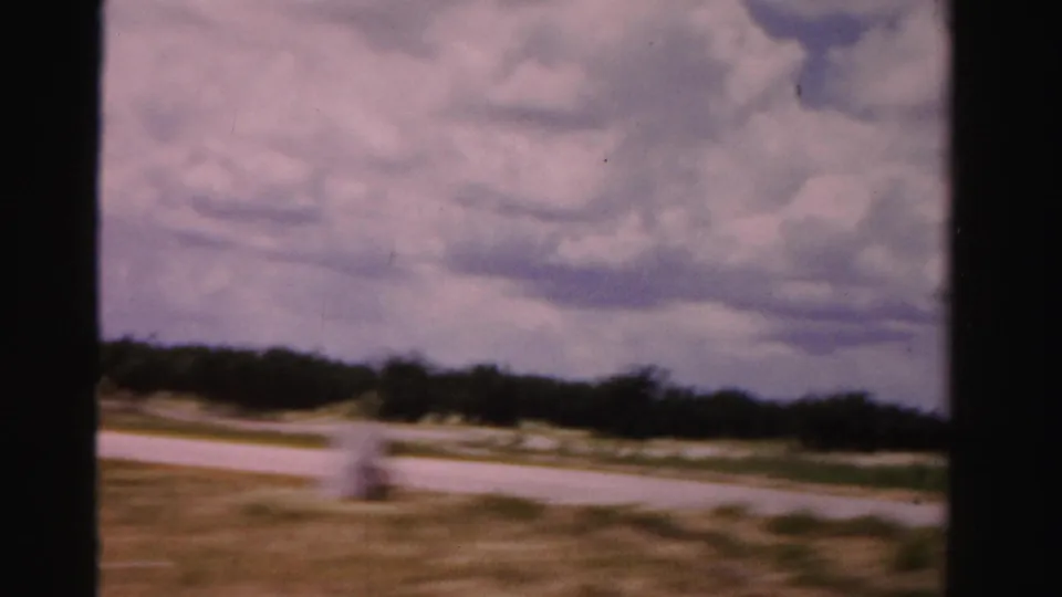 1955 Dade County road-trip view of an open grassy roadside, pale road curve, distant tree line, and cloud-filled sky from a moving car.