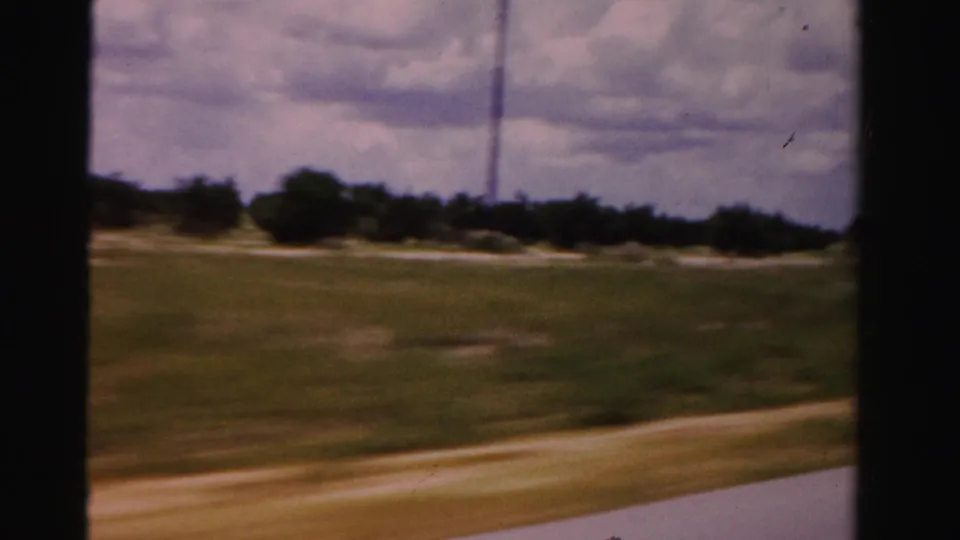 1955 Dade County moving-car frame showing grass, a road edge, distant trees, a slim tower or pole, and a cloudy sky.
