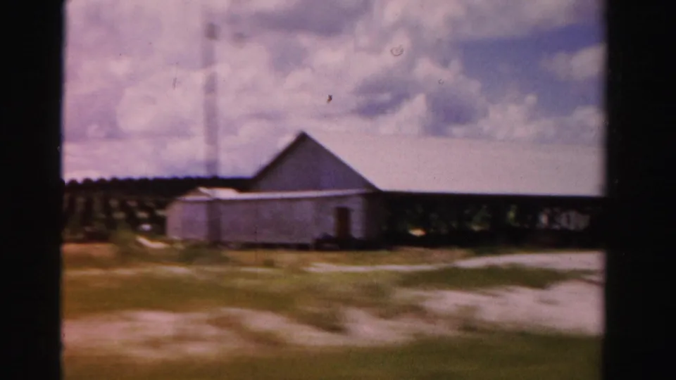 Rural Dade County scene with low farm structures, a tall tower, open ground, and cloud cover in a moving 8mm shot.