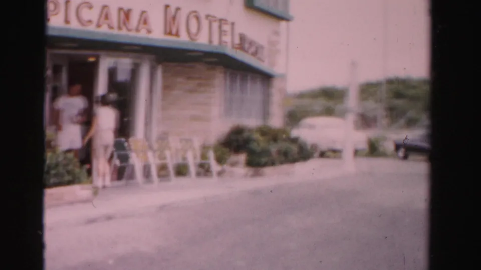 1955 Florida motel stop showing a partial "ICANA MOTEL" sign, two adults at the entrance, outdoor chairs, shrubs, and cars.