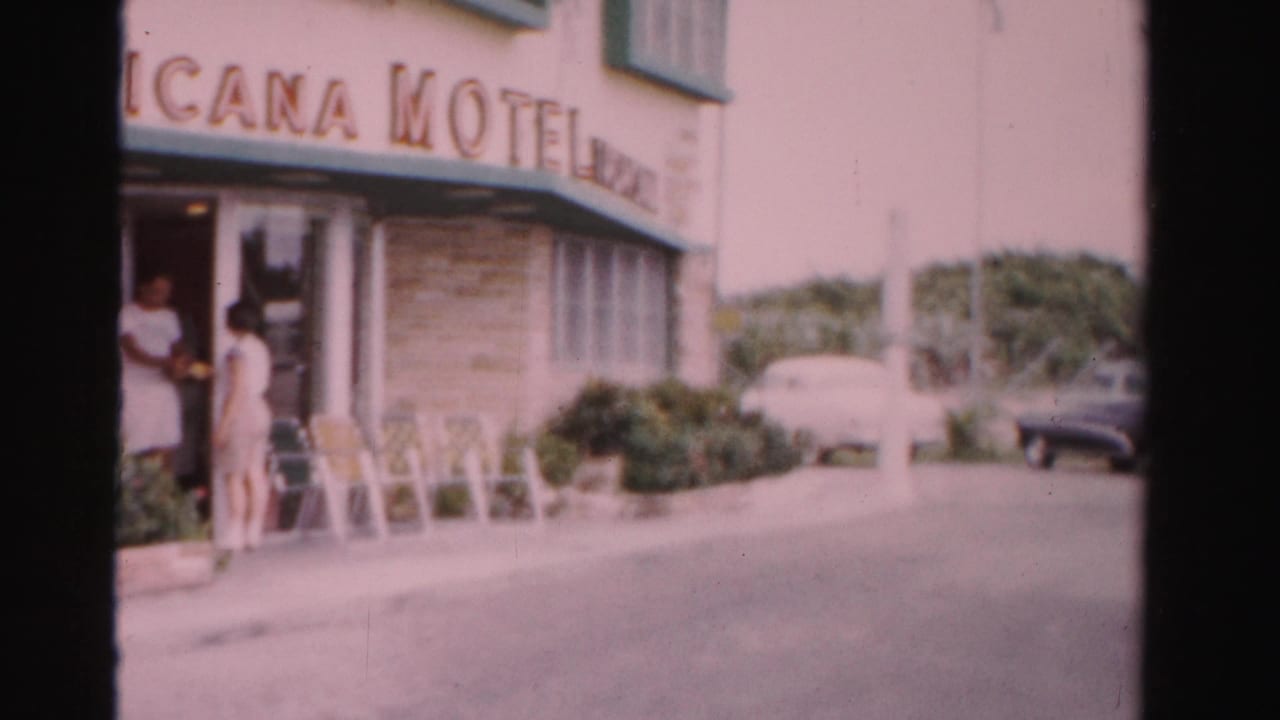 Woman near a motel entrance with a Tropicana Motel sign, patio chairs, shrubs, and parked cars.