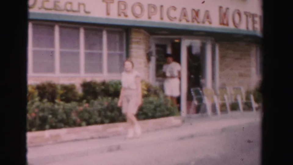 1955 Dade County motel exterior with a woman walking past windows, shrubs, entrance chairs, and Tropicana Motel signage.