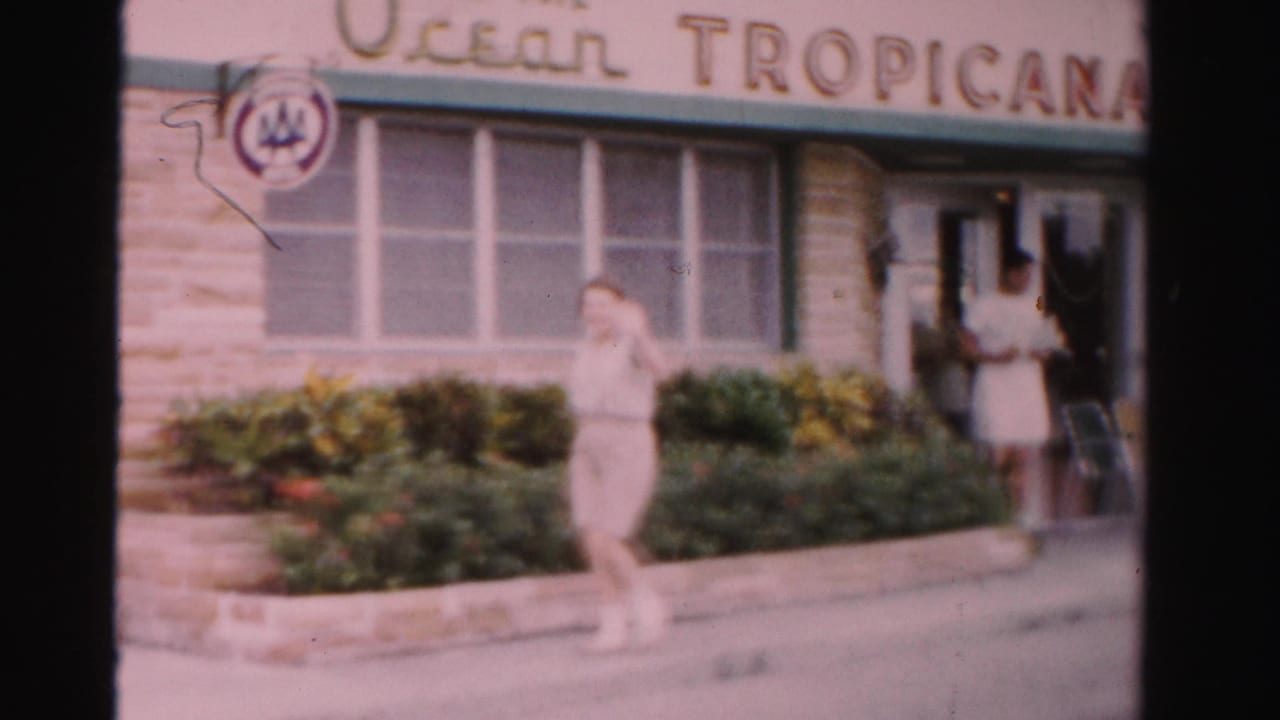 Woman walking past the Ocean Tropicana Motel entrance with shrubs and signage visible.