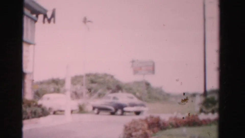 The final frame looks across a motel-side parking area with vintage cars, grass, a road edge, and a distant sign in 1950s Dade County footage.