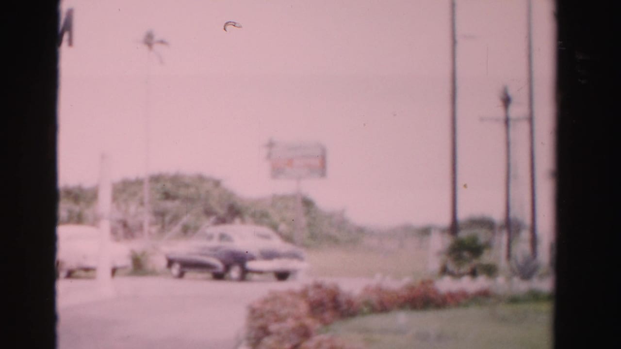 Curved roadside with parked mid-century cars, hedges, utility poles, and distant signs.