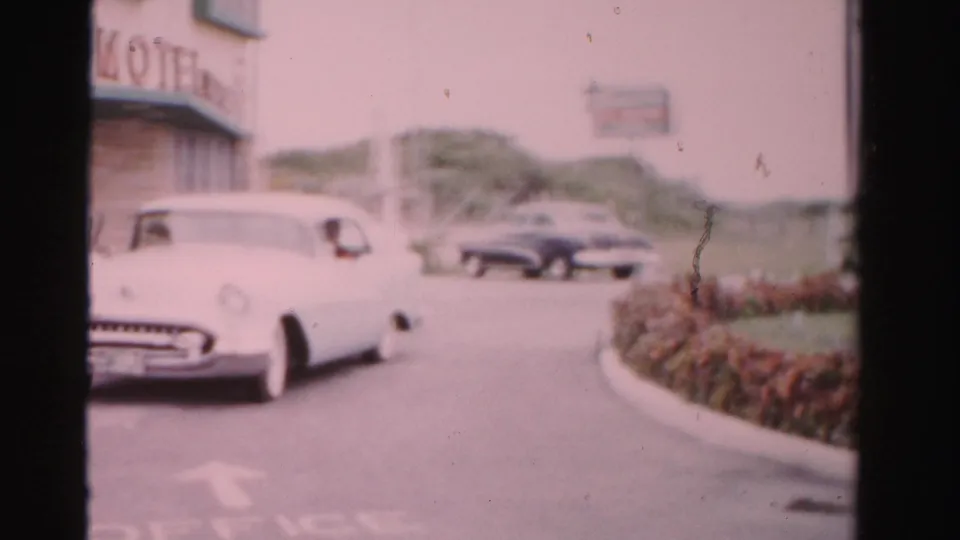 White 1950s car passes near a low roadside building in Dade County, with hedges and another car beyond.