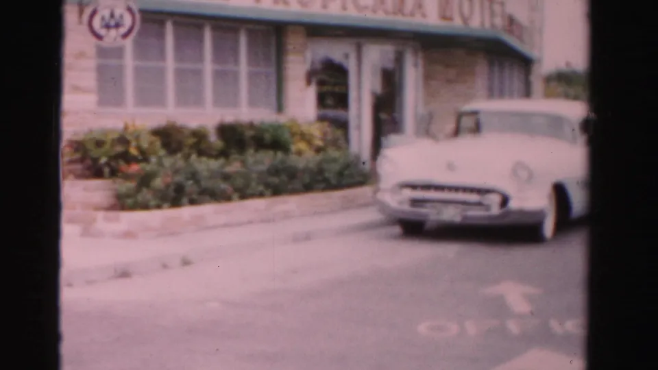 1955 Dade County frame with a white car outside a low roadside building with green awning, windows, shrubs, and curb.
