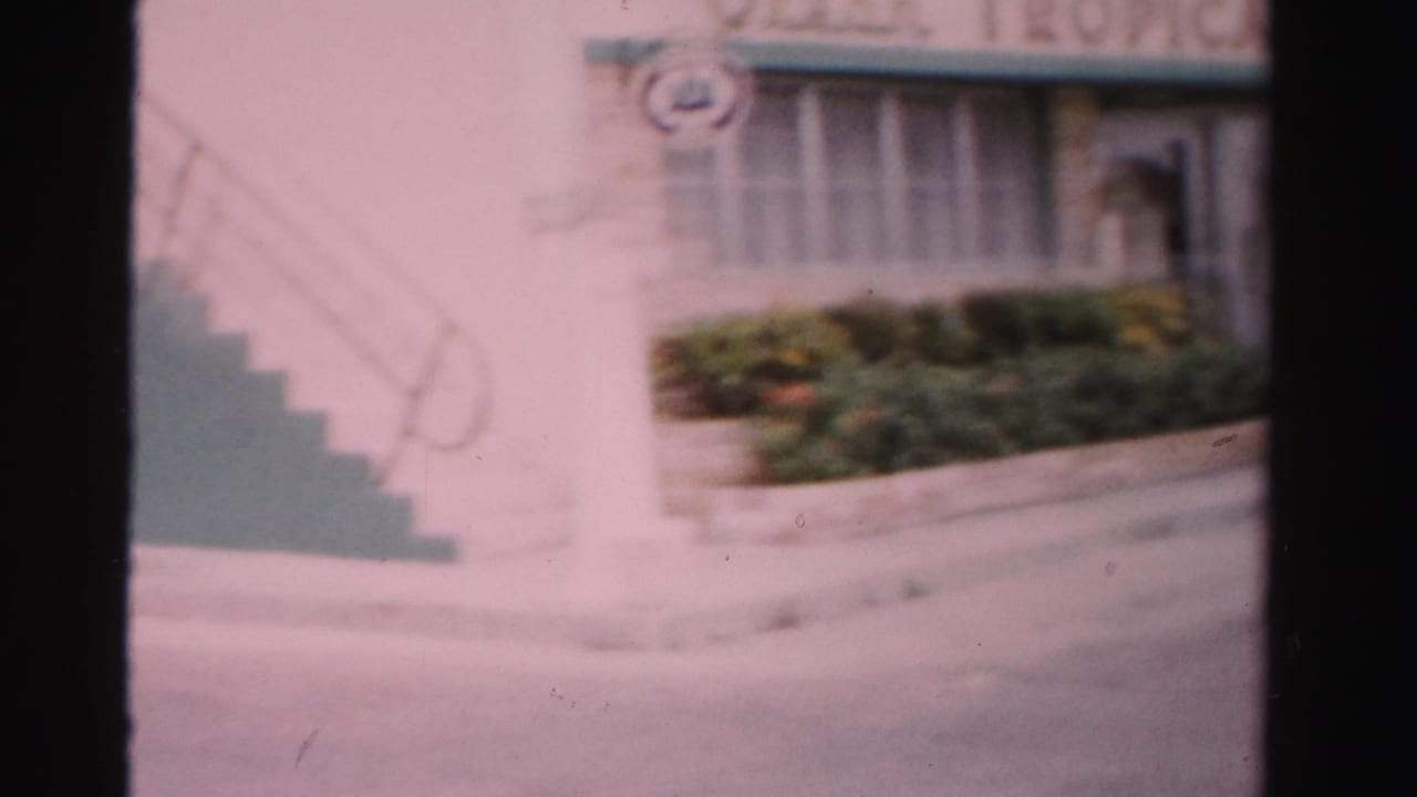 Motel frontage with green trim, exterior stairs, shrubs, and partial sign above the entrance.