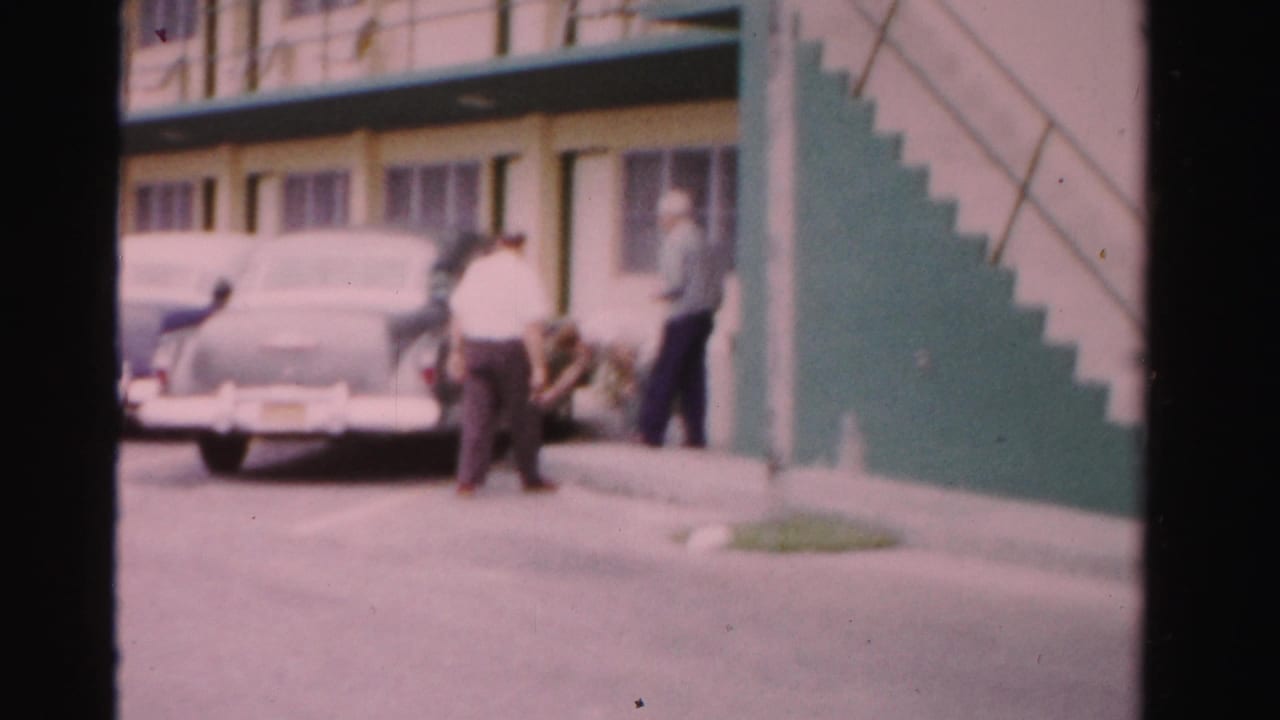 Adults stand near parked mid-century cars outside a motel with green trim.