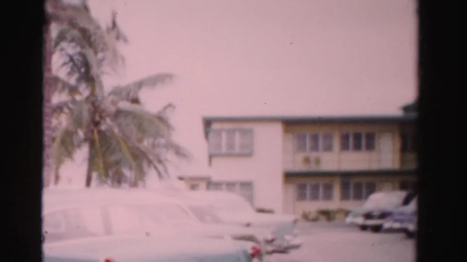 1955 Florida home-movie frame with palm trees, parked cars, and a pale two-story motel-like exterior.