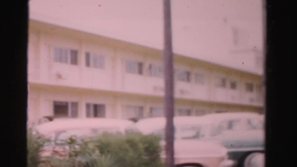 Soft 8mm view of a white Dade County building with palm trunk, shrubs, and parked cars, consistent with the 1955 clip metadata.