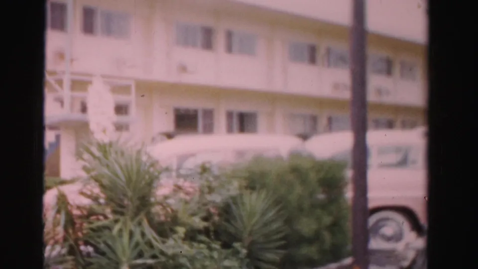1955 Dade County home-movie frame with palm landscaping, parked cars, and a white residential or motel-like building beyond.