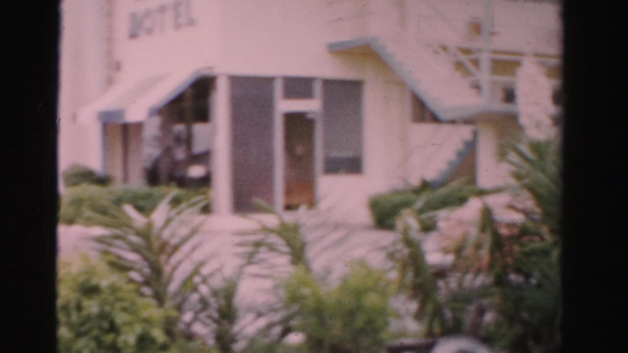1955 Dade County hotel entrance area with a partially readable sign, awning, exterior stairway, landscaped shrubs, and nearby parked cars.