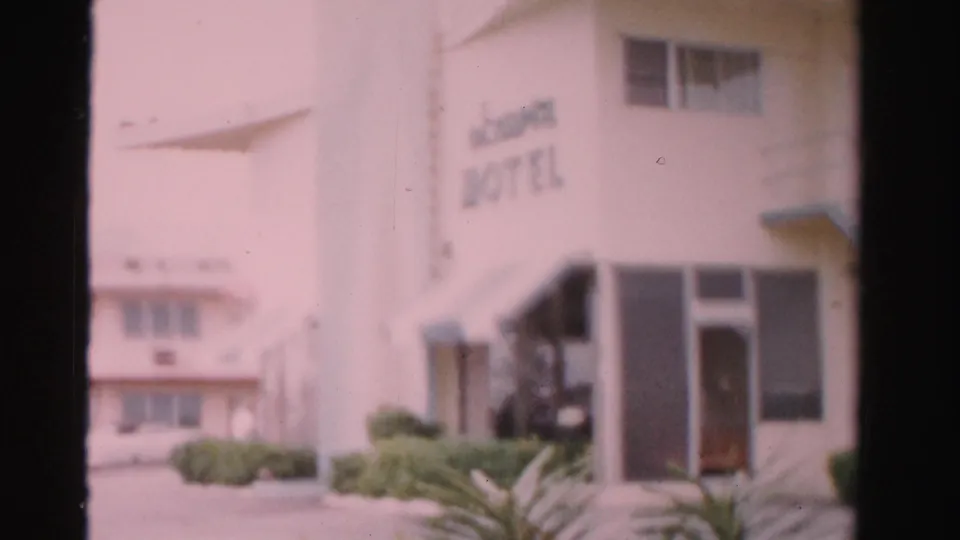 Soft 8mm view of a Dade County hotel facade in 1955, showing entrance doors, partial sign lettering, windows, landscaping, and a parked car.
