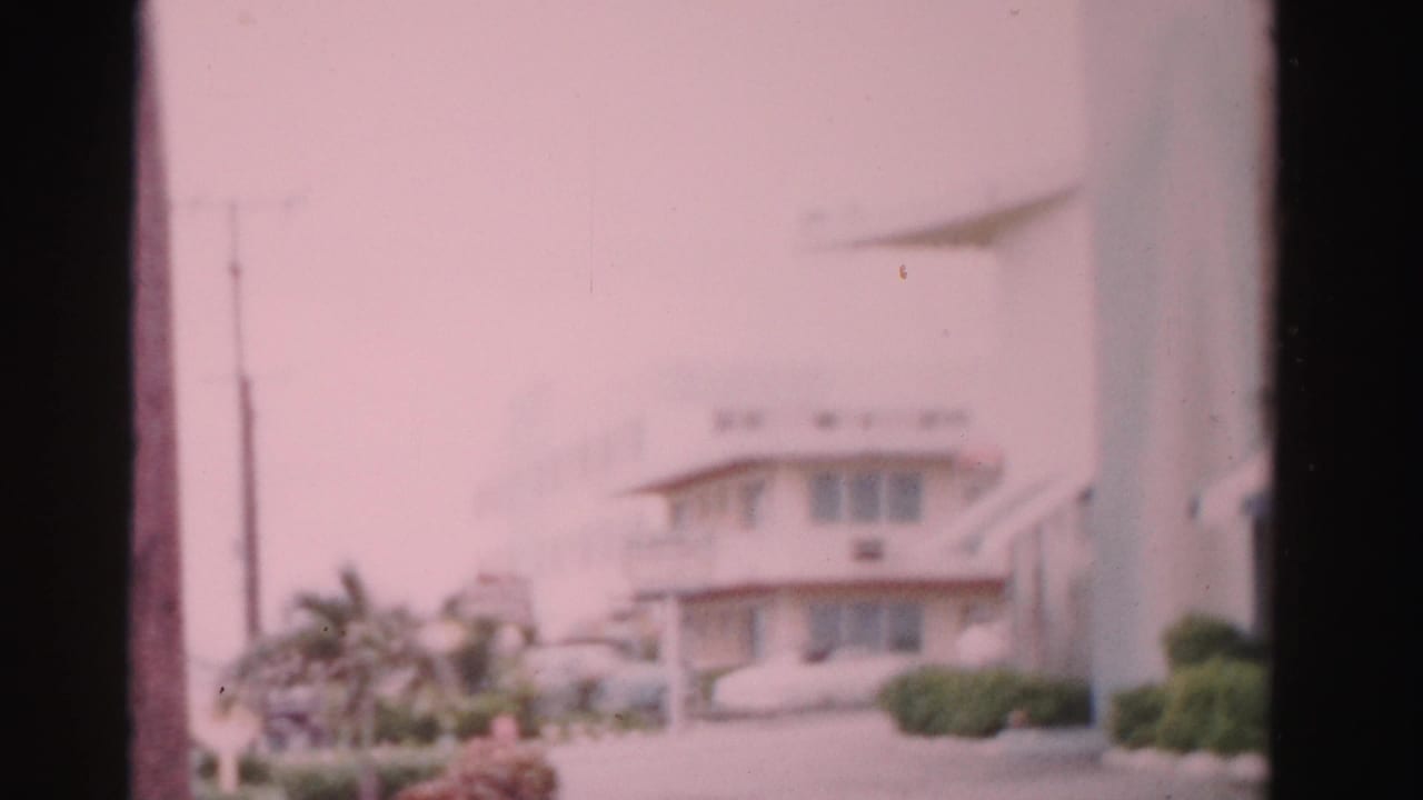 Soft 8mm frame of a pale hotel building with palm landscaping and a parked car nearby.