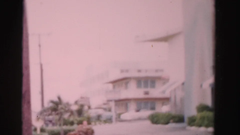 1955 Dade County home-movie frame showing a pale hotel exterior, palm trees, low shrubs, and a parked car in soft focus.