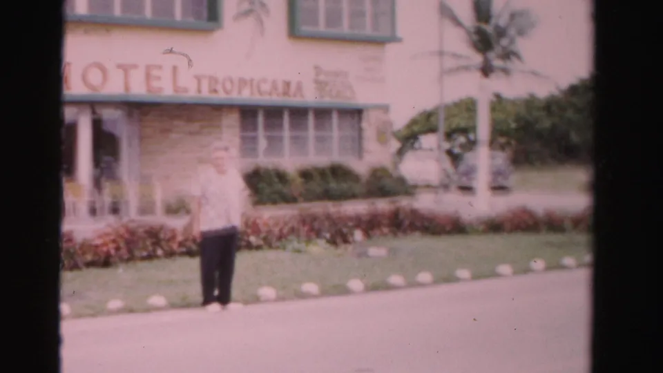 1955 Dade County frame of an adult man outside Hotel Tropicana, with the sign, palm trees, hedges, and hotel entrance visible.