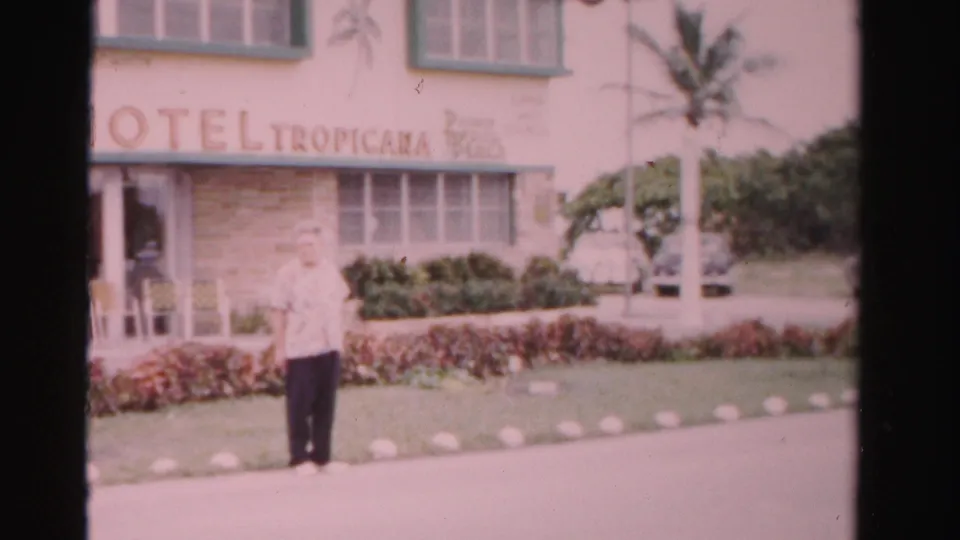 1955 Dade County home-movie frame showing an adult man outside Hotel Tropicana, with a visible sign, palm tree, hedges, and lawn.