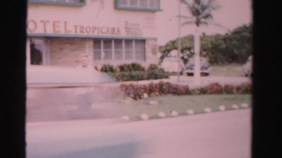 1955 Dade County frame of the Hotel Tropicana exterior, showing the sign, palm landscaping, trimmed hedge, sidewalk, and a partial figure.