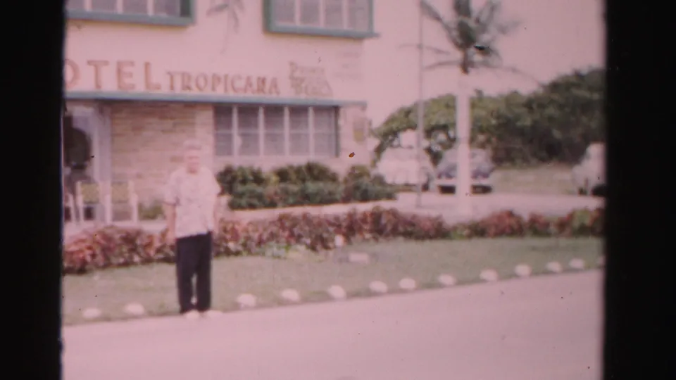 1955 Dade County home-movie frame showing a man near a Tropicana-signed building with palms, hedges, and cars behind him.