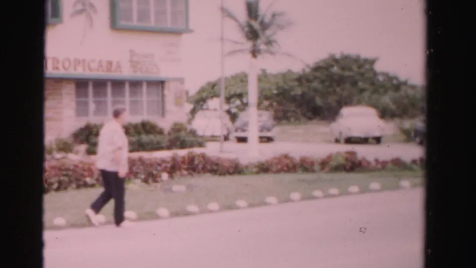 1955 Dade County frame of a man walking along a paved roadside past Tropicana signage, palms, hedges, and parked cars.