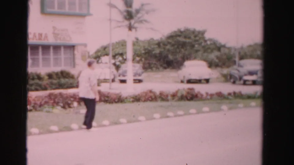 1955 Dade County home-movie frame showing the man farther along the roadside with palms, hedges, and parked cars nearby.