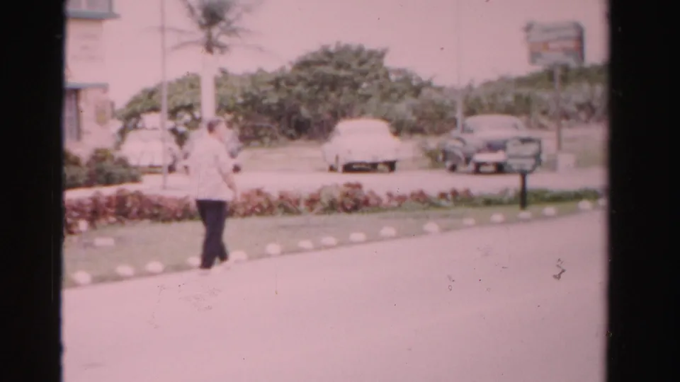 1955 Dade County frame showing a man walking past hedges and parked cars, with palms and a distant roadside sign visible.