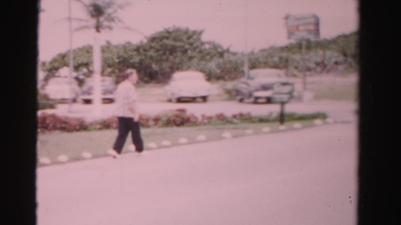 Older man walking beside a landscaped street with parked cars and palms in the background.