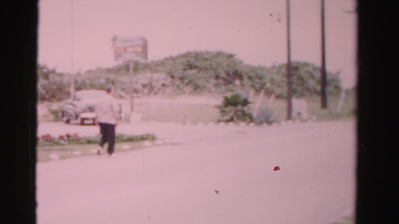 1955 Dade County 8mm frame showing a man walking beside a road with mid-century cars, low shrubs, utility poles, and a roadside sign.