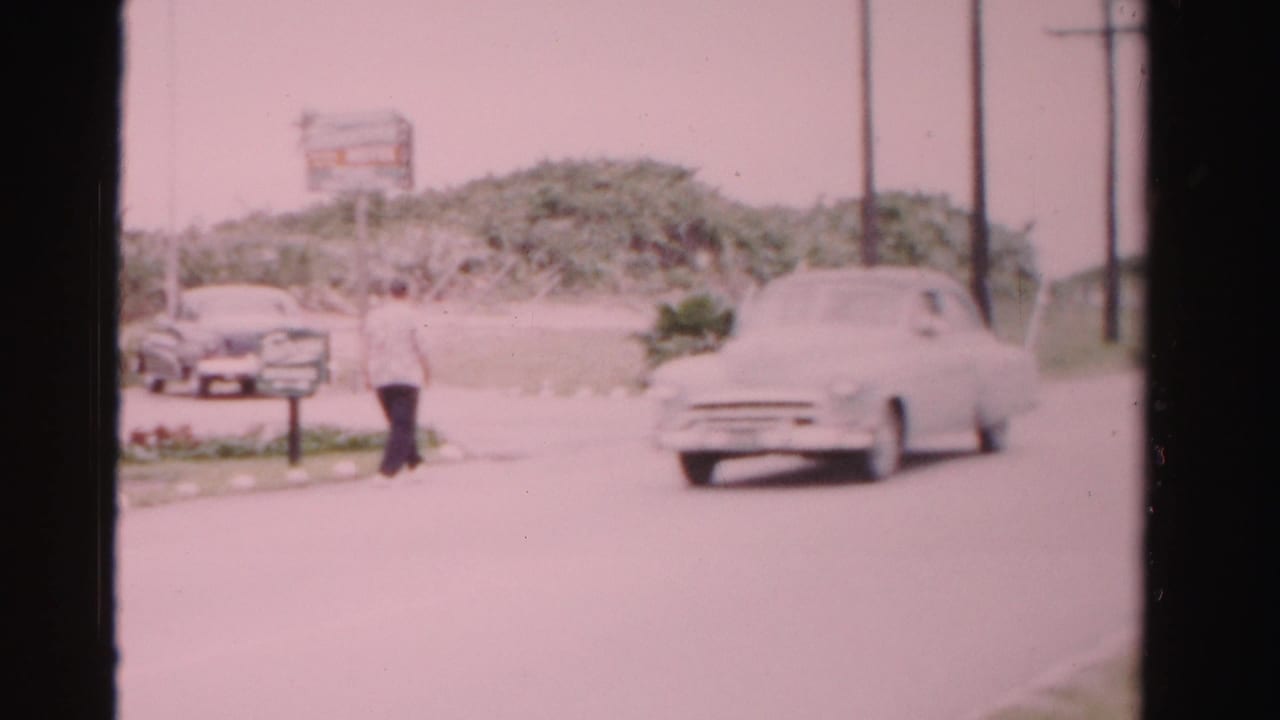 A white mid-century sedan passes through a Dade County roadside frame while a man walks near parked cars, shrubs, poles, and a sign.