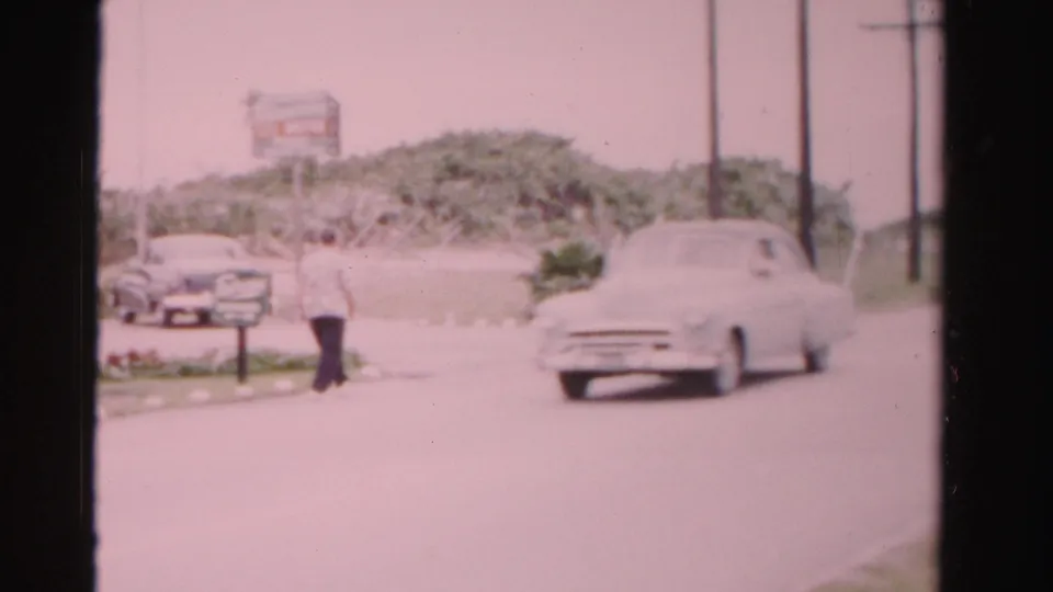 A white mid-century sedan passes through a Dade County roadside frame while a man walks near parked cars, shrubs, poles, and a sign.