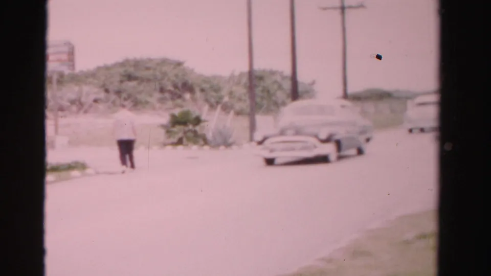 Soft 8mm frame from 1955 Dade County with a passing classic sedan, distant pedestrian, roadside plants, utility poles, and hazy sky.