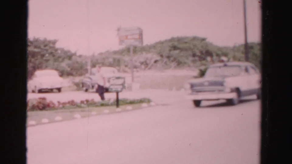 Dade County roadside scene with a man near parked cars, a passing sedan, low tropical plantings, a sign, and visible 8mm film borders.