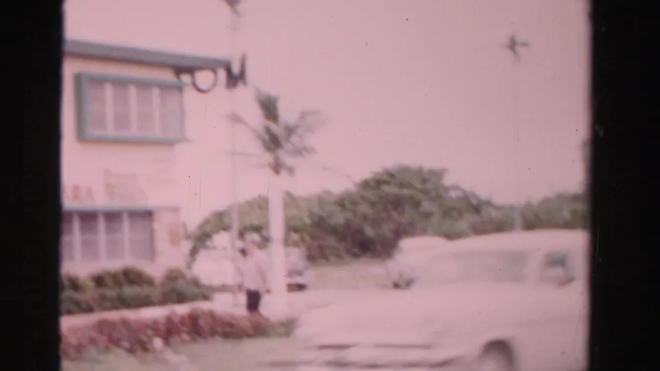 1955 Dade County frame showing part of a motel building, a palm tree, shrubs, and a blurred car passing in the foreground.