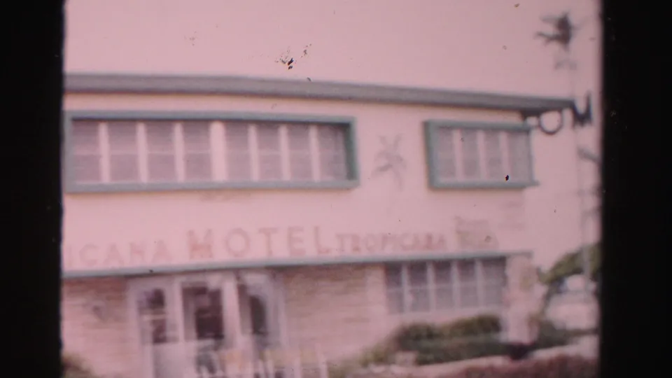 1955 Dade County home-movie frame showing the Tropicana Motel facade, entrance doors, upper windows, shrubs, and a partial car.