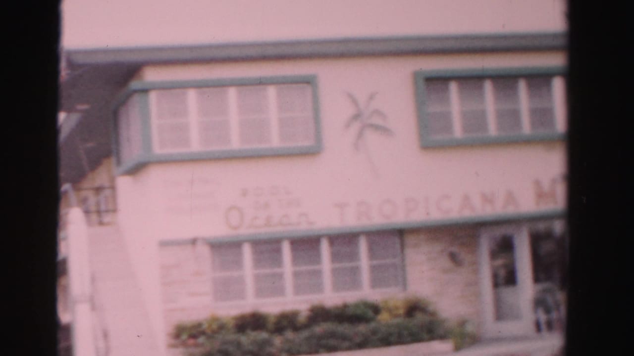Closer motel exterior with painted signage, windows, shrubs, and a doorway at right.