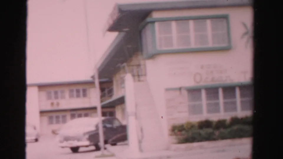 Angled 1955 motel view with green-trimmed upper window, partial Tropicana Motel signage, balcony railings, shrubs, and parked cars.