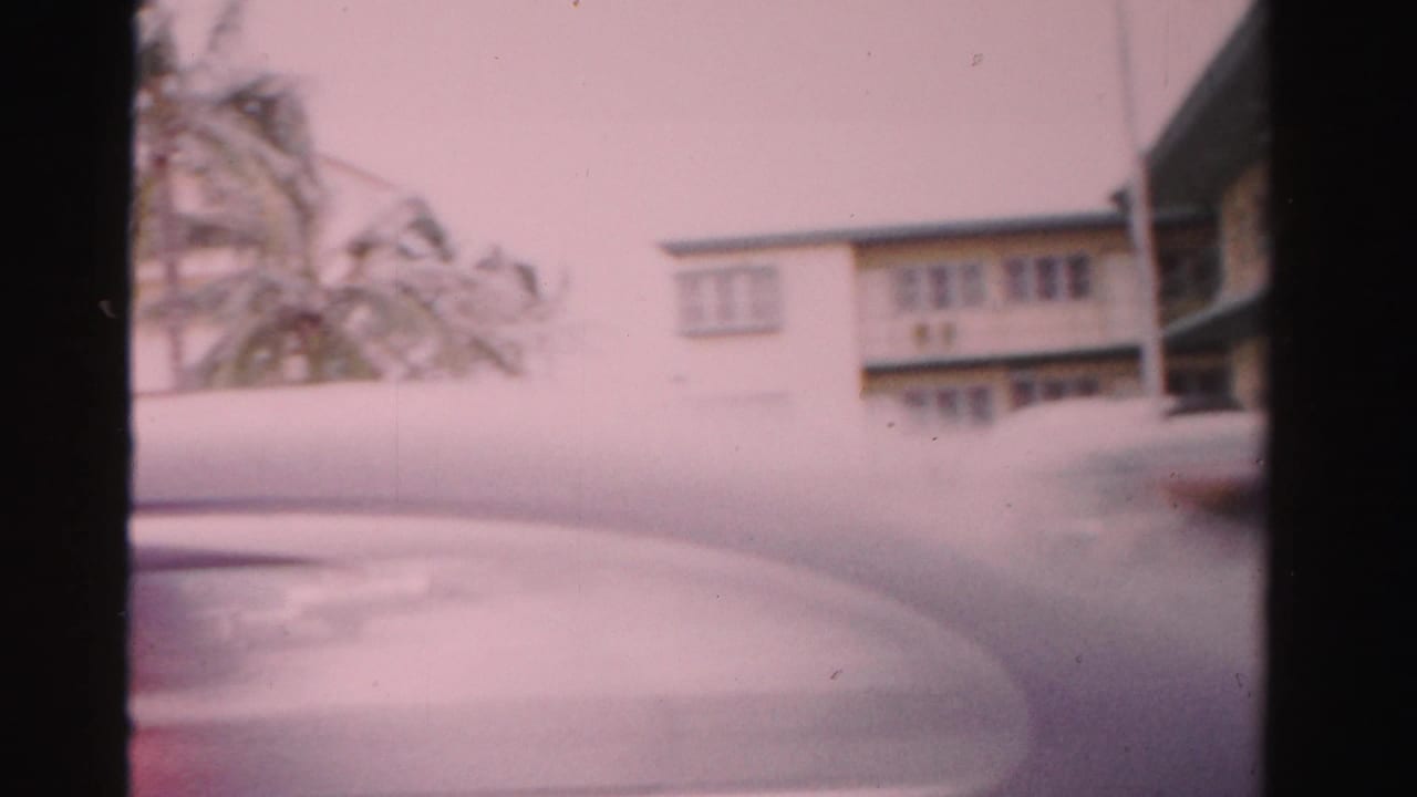 Soft 8mm frame of a motel frontage partly obscured by a passing car and film gate edges.