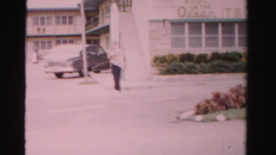 1955 Dade County home-movie frame with an adult man walking beside the Ocean Tropicana Hotel frontage and parked mid-century cars.