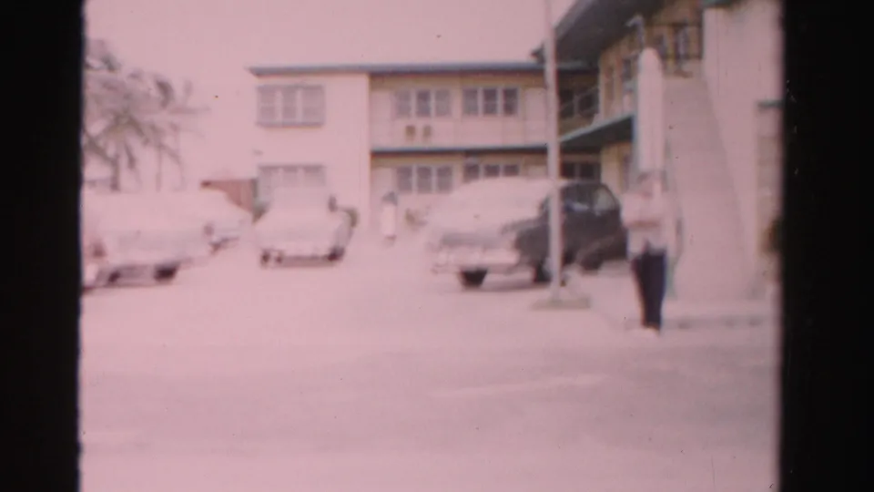 Wide 1955 Dade County 8mm view of parked mid-century cars outside a motel, with a streetlight, palm trees and an adult man near the right edge.